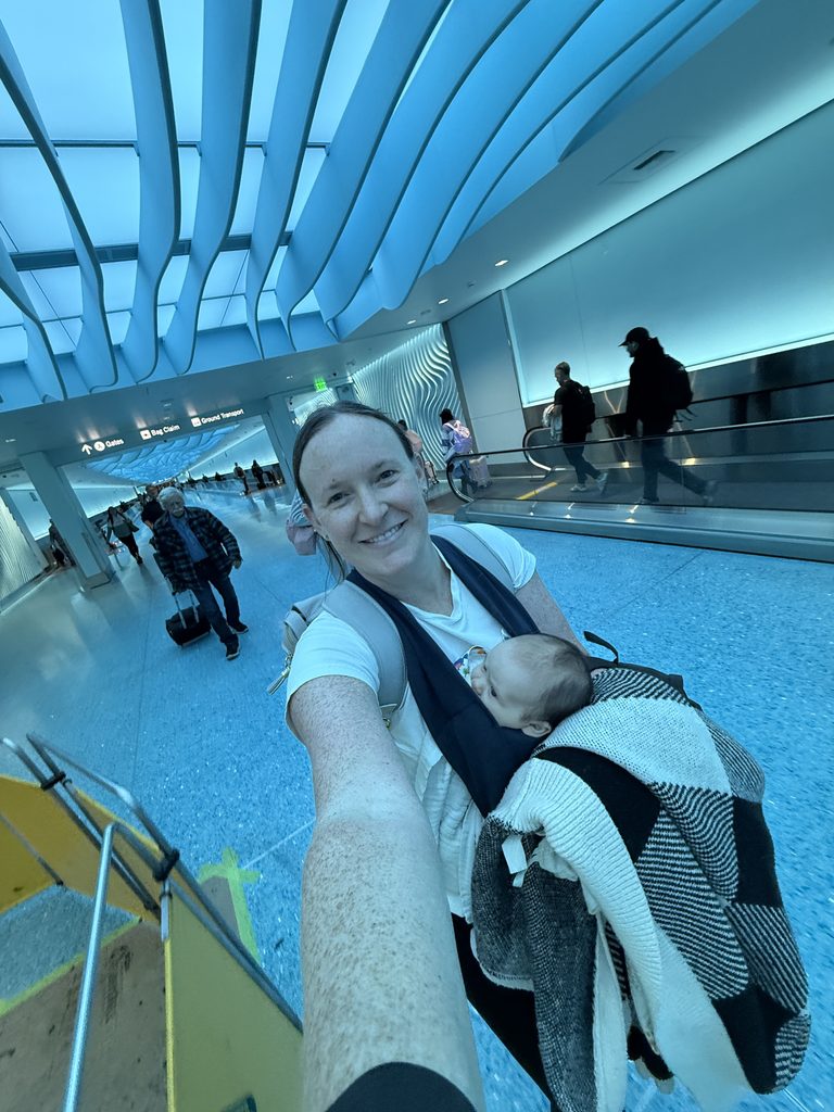 A mother takes a selfie while wearing her 9-week-old baby in a navy blue baby carrier at the airport. She smiles at the camera as travelers walk through the terminal behind her.