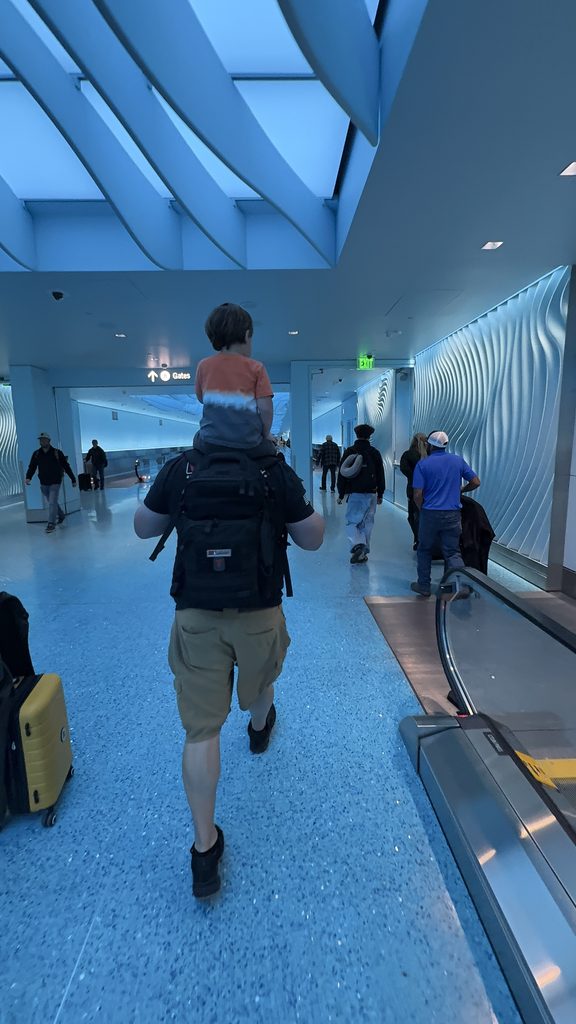 A father walks through the airport carrying his toddler on his shoulders. The toddler wears an orange shirt and looks ahead toward the gates as other travelers move through the terminal.