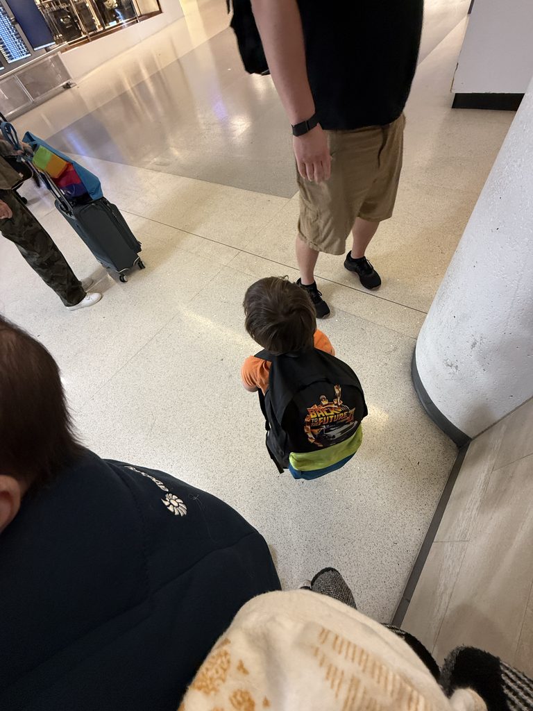 A toddler wearing an orange shirt and a colorful Back to the Future backpack walks through the airport beside his parents. The photo is taken from above, showing the child carrying his own bag.