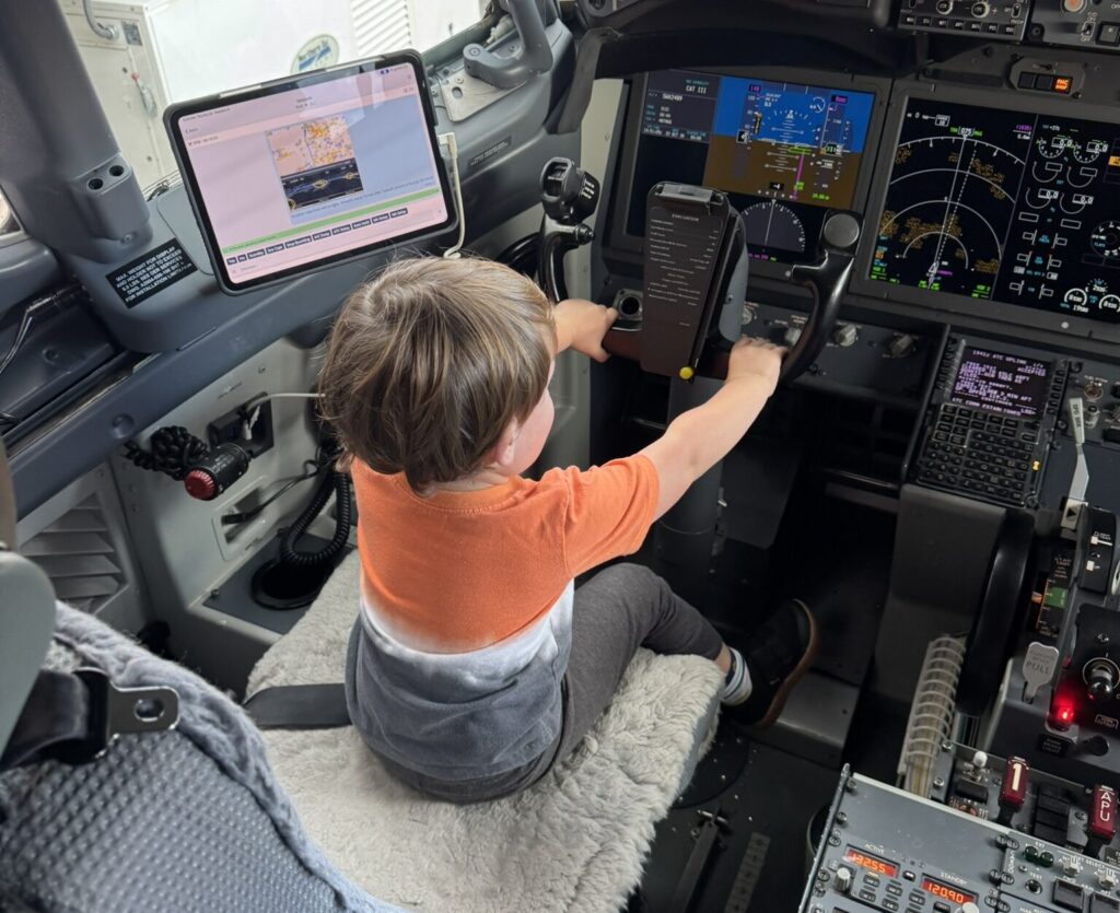 A toddler in an orange shirt sits in an airplane cockpit, holding the controls and looking at the instrument panels. Flight displays, buttons, and screens surround the child.